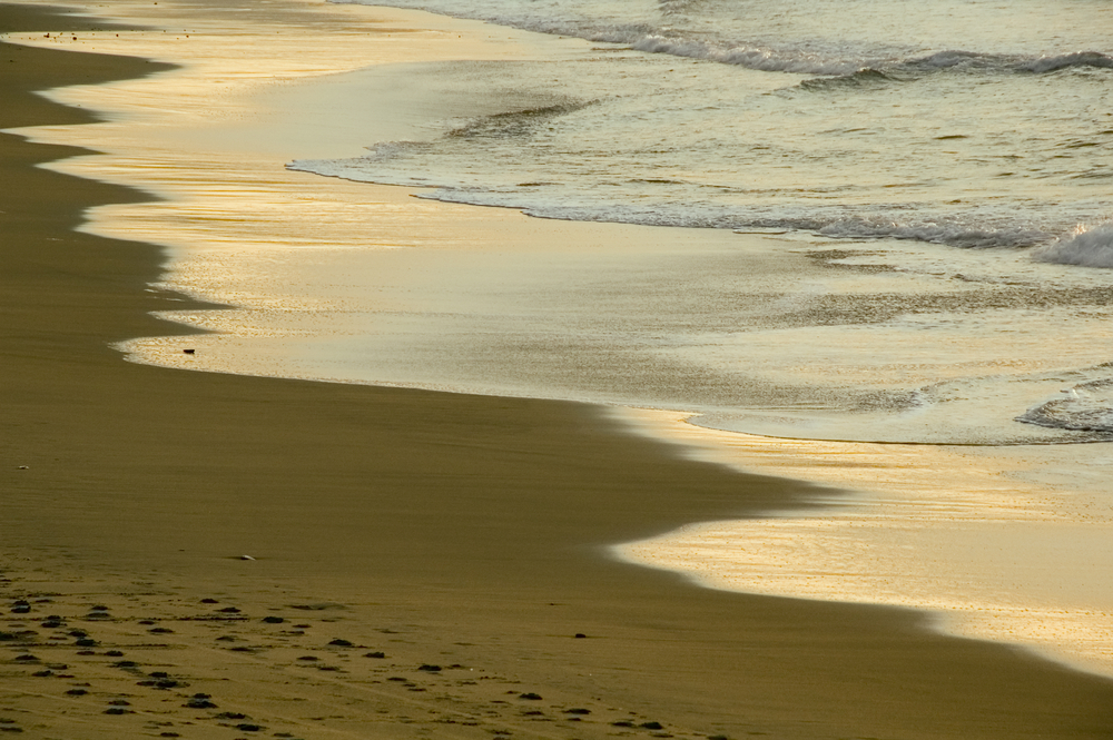 Ocean waves reflect light of setting sun on sandy beach with footprints